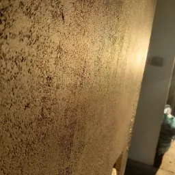 Ściana z grubą, ziarnistą fakturą w ciepłym świetle słonecznym, widoczny fragment narożnika i podłogi, w tle worek z odpadami budowlanymi.