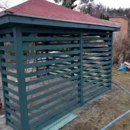 Drewniana altana pomalowana na zielono z czerwonym dachem z gontu bitumicznego, widoczna w ogrodzie na tle drzew bez liści.