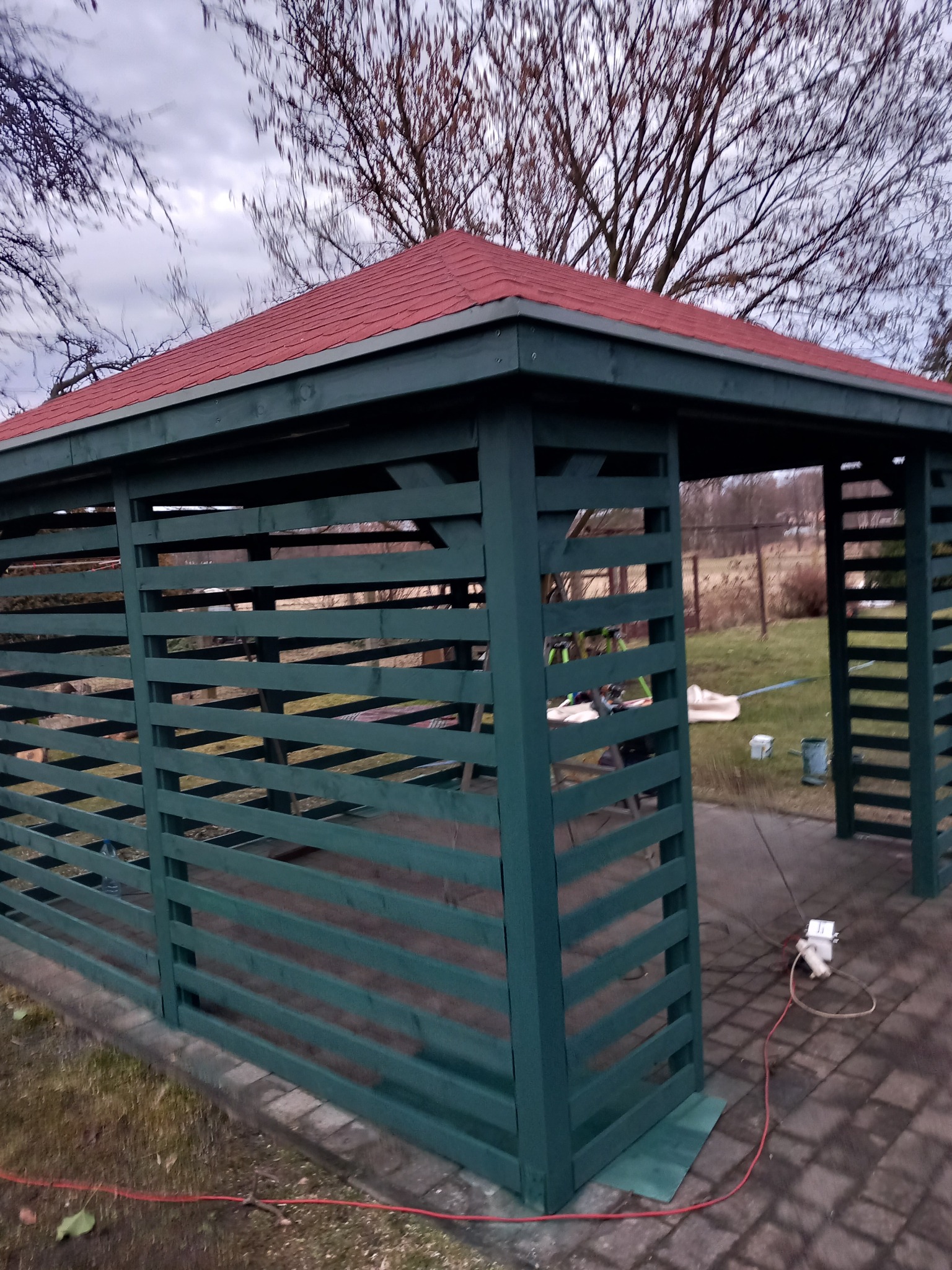 Zielona altana z czerwonym dachem, częściowo osłonięta poziomymi listwami, na kostce brukowej, z widocznymi narzędziami i puszkami farby wewnątrz, sugerującymi świeże malowanie.