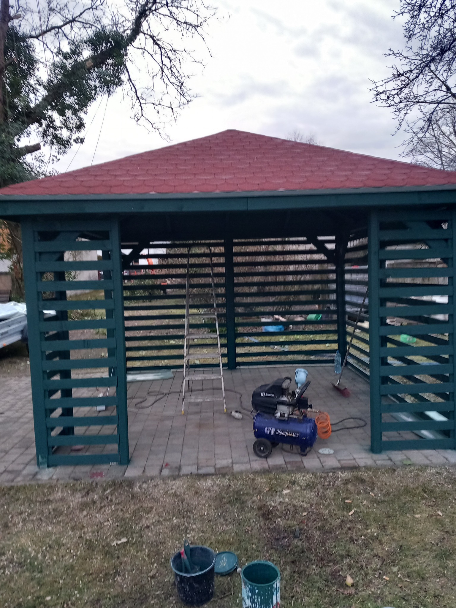 Zielona, drewniana altana z czerwoną dachówką na kostce brukowej, widoczny kompresor, drabina i wiaderka z farbą, sugerujące etap budowy lub wykończenia.