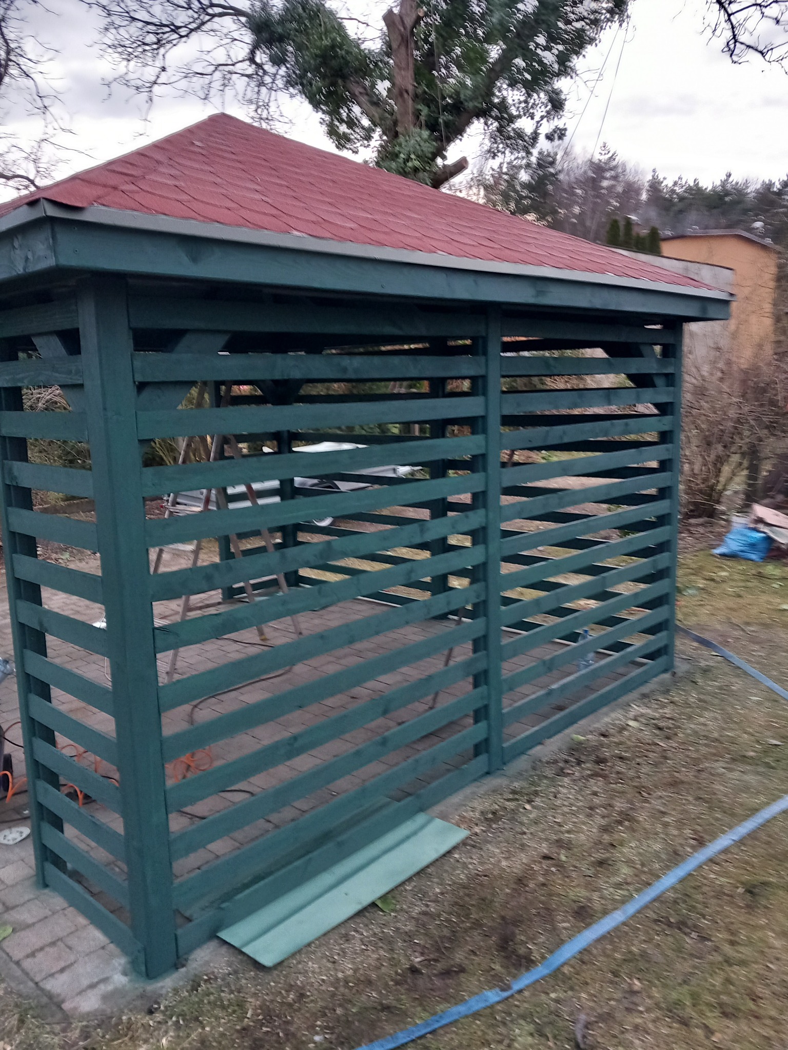 Drewniana altana pomalowana na zielono z czerwonym dachem z gontu bitumicznego, widoczna w ogrodzie na tle drzew bez liści.