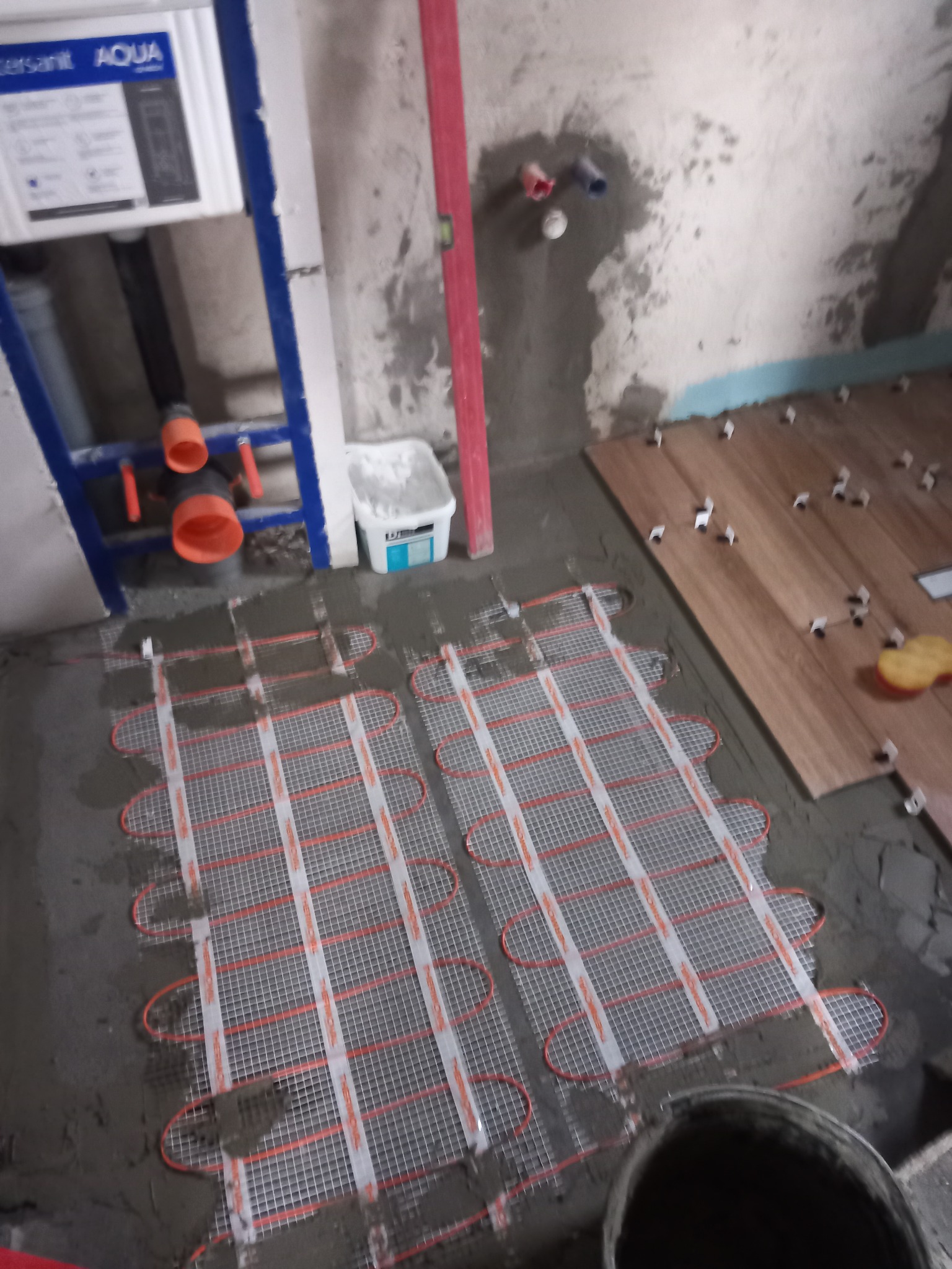 Montaż ogrzewania podłogowego w łazience, widoczne maty grzewcze zatopione w wylewce, częściowo ułożone płytki imitujące drewno oraz instalacja podtynkowa WC.