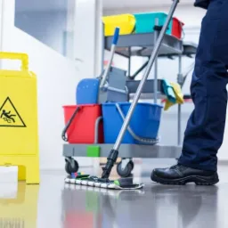 Pracownik w granatowym uniformie myje podłogę mopem w biurze, obok żółty znak ostrzegawczy i wózek z akcesoriami do sprzątania.