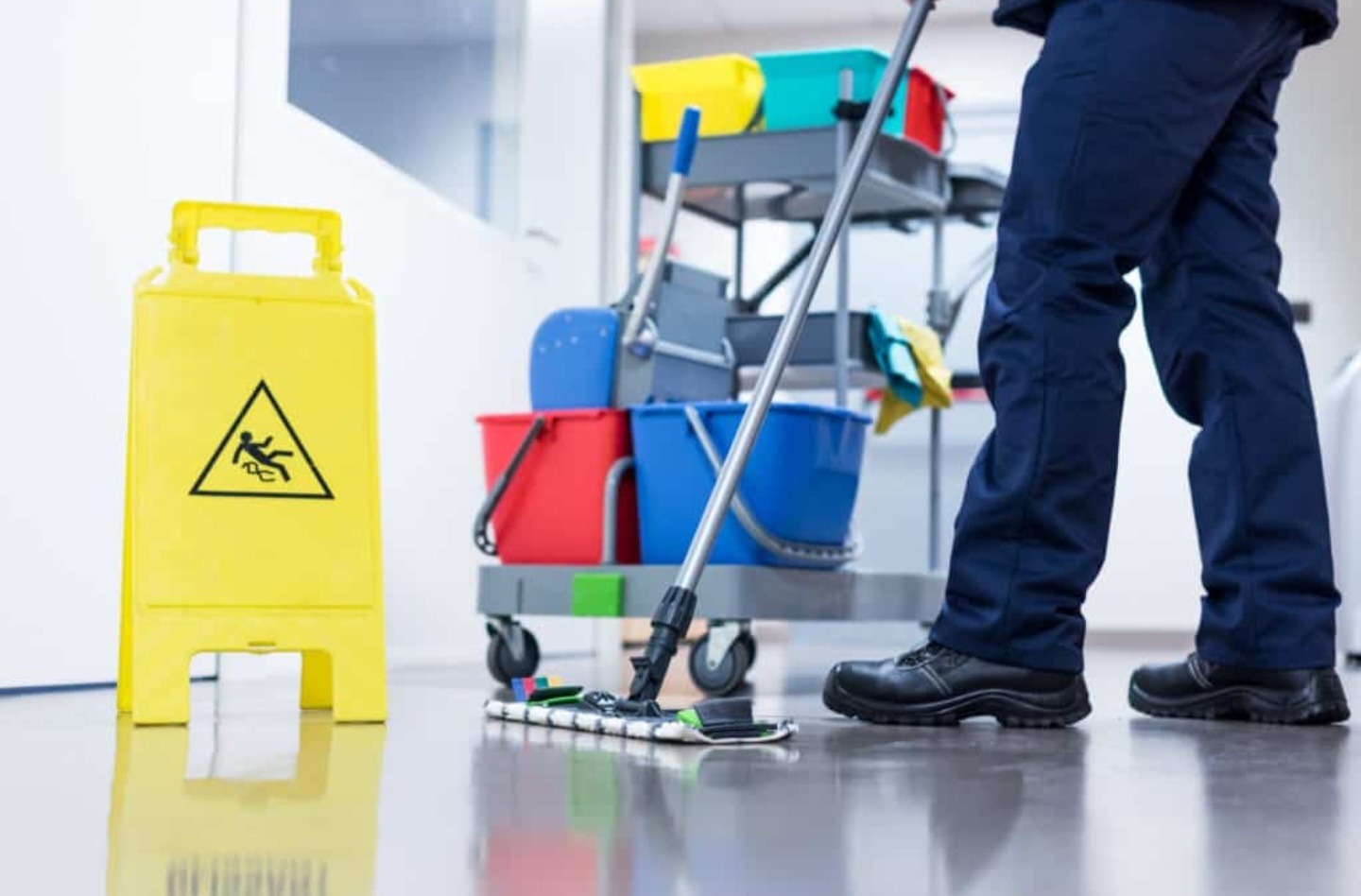 Pracownik w granatowym uniformie myje podłogę mopem w biurze, obok żółty znak ostrzegawczy i wózek z akcesoriami do sprzątania.