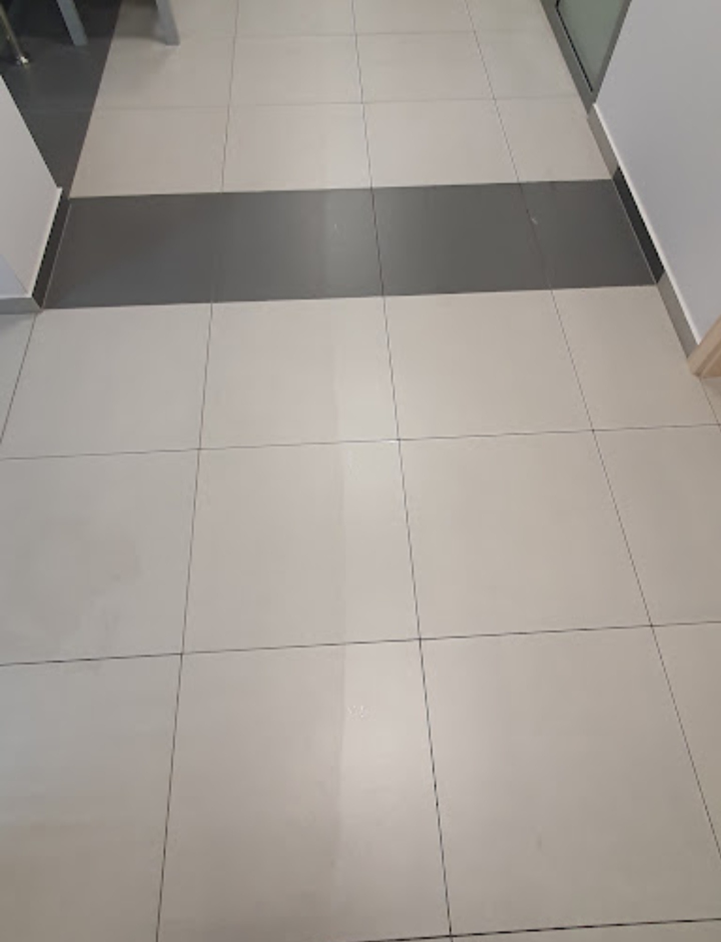 Wyraźnie widoczna różnica w czystości pomiędzy umytą a nieumytą częścią jasnobeżowej podłogi z płytek, z szarym pasem płytek w środku.