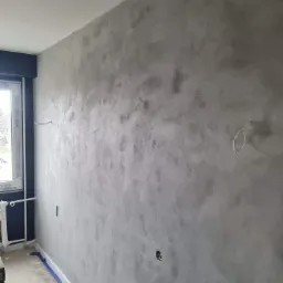 Ściana z efektem dekoracyjnym w odcieniach szarości, zabezpieczona listwa przypodłogowa taśmą malarską, widoczne wystające przewody elektryczne w Zgierzu.