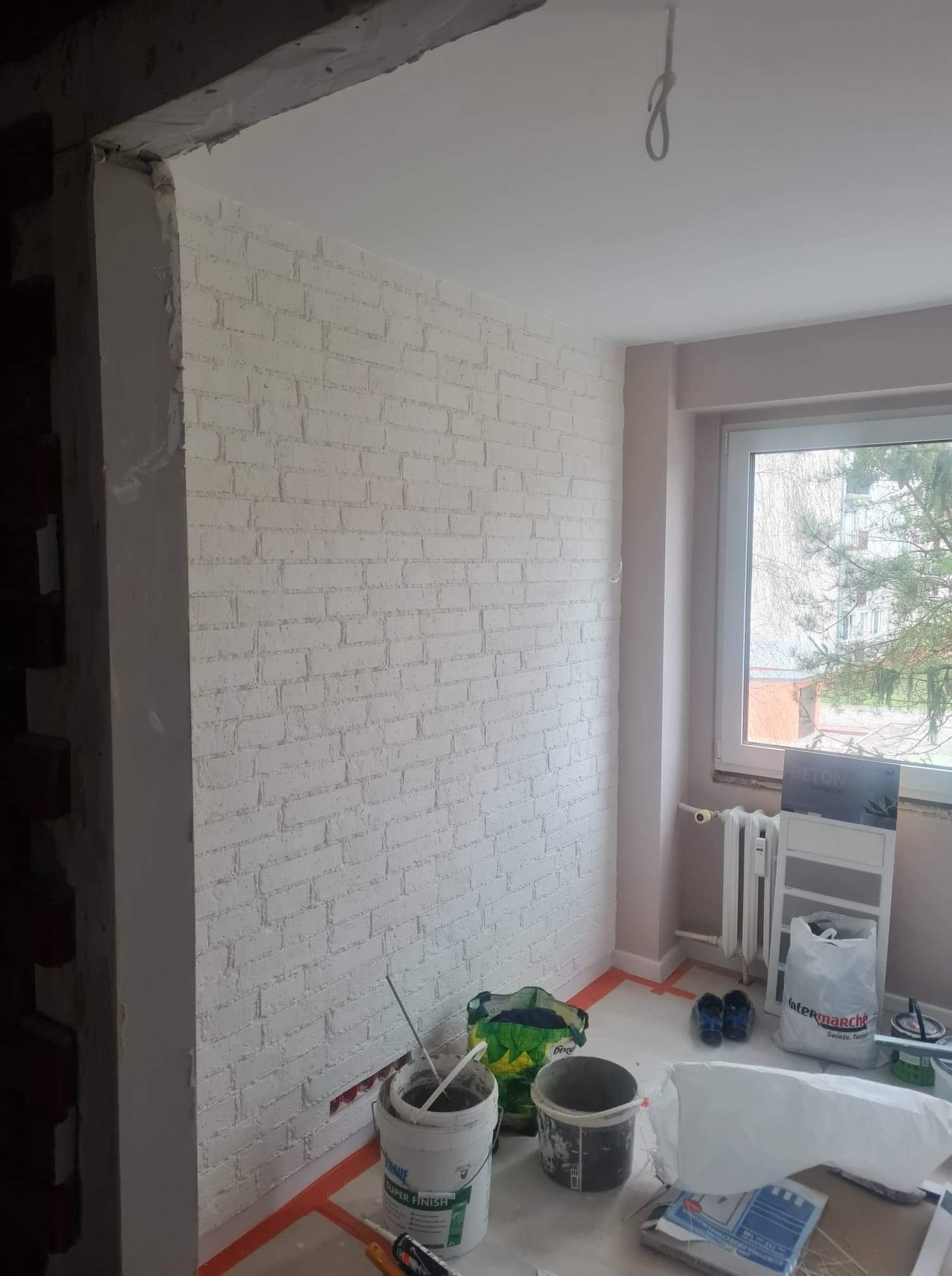 Ściana z cegły pomalowana na biało z widocznymi narzędziami i materiałami budowlanymi podczas prac wykończeniowych, zabezpieczona pomarańczową taśmą, w tle okno z widokiem na zewnątrz.