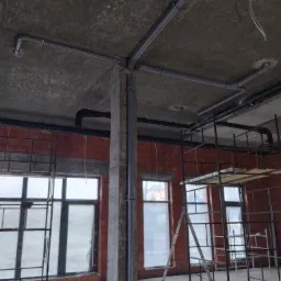 Szare rury kanalizacyjne przymocowane do sufitu w surowym stanie, widoczne rusztowania i okna w tle, wnętrze w trakcie remontu.