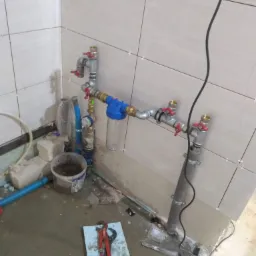 Nowe przyłącze wodne z filtrem i zaworami, widoczne narzędzia na podłodze w trakcie montażu, płytki na ścianach z krzyżykami dystansowymi.