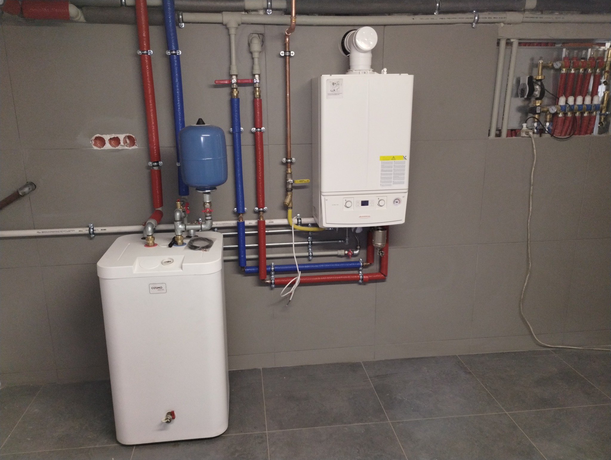 Kotłownia z białym piecem gazowym, zasobnikiem CO marki Cosmo, niebieskim naczyniem przeponowym i instalacją z rur miedzianych i plastikowych w kolorach czerwonym i niebieskim, na tle szarych...