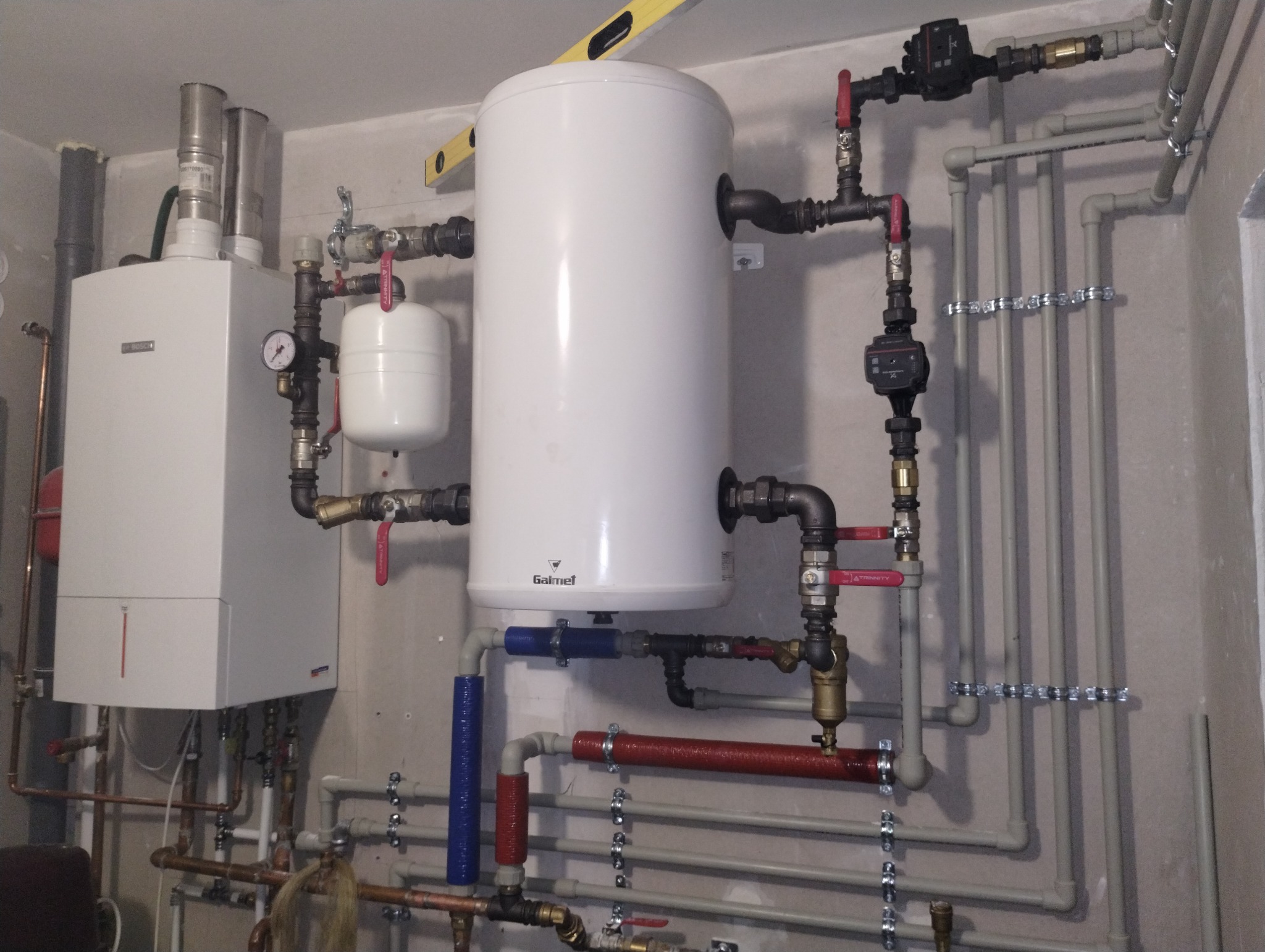 Instalacja grzewcza w kotłowni z białym zasobnikiem ciepłej wody marki Galmet, piecem gazowym Bosch, manometrem, zaworami z czerwonymi rączkami i rurami w izolacji termicznej, poziomica na górze...
