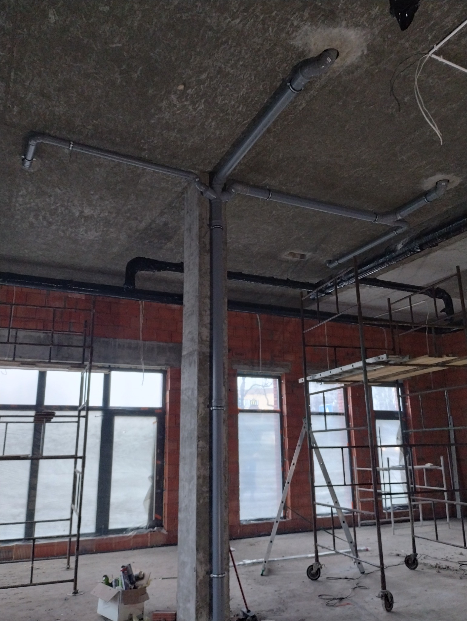 Szare rury kanalizacyjne przymocowane do sufitu w surowym stanie, widoczne rusztowania i okna w tle, wnętrze w trakcie remontu.