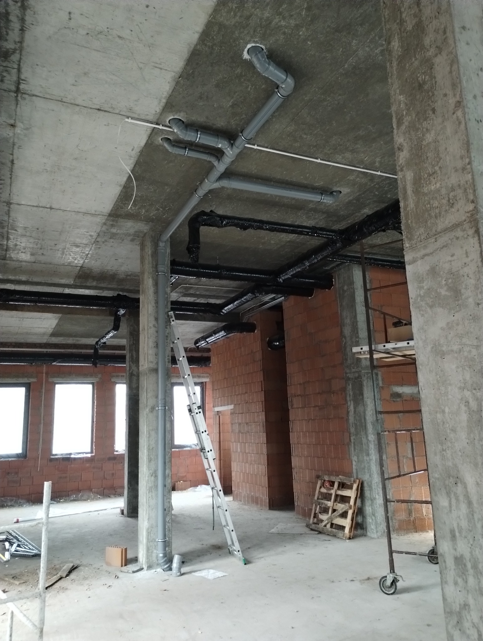 Surowy stan budynku w trakcie instalacji: szare rury kanalizacyjne i czarne rury grzewcze pod betonowym sufitem, drabina oparta o słup, ściany z cegieł.