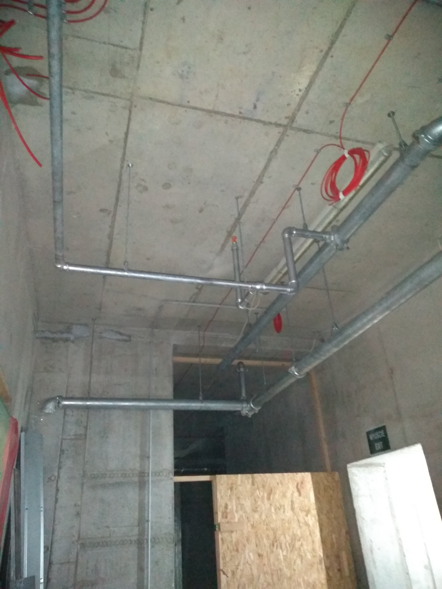 Szare metalowe rury pod betonowym sufitem w surowym wnętrzu budynku, widoczne czerwone kable i fragment ściany z otwartym przejściem.