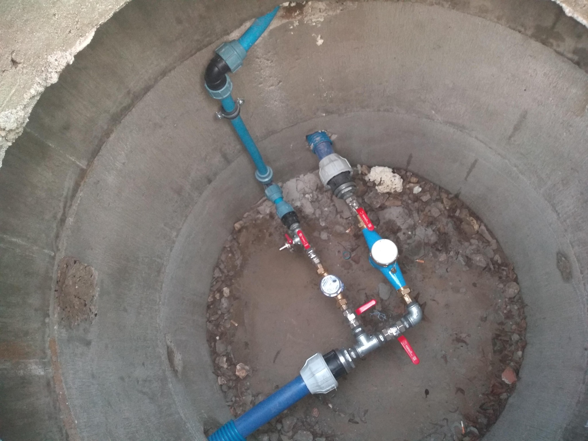 Studzienka betonowa z niebieskimi rurami wodociągowymi, zaworami i wodomierzami, widok z góry.