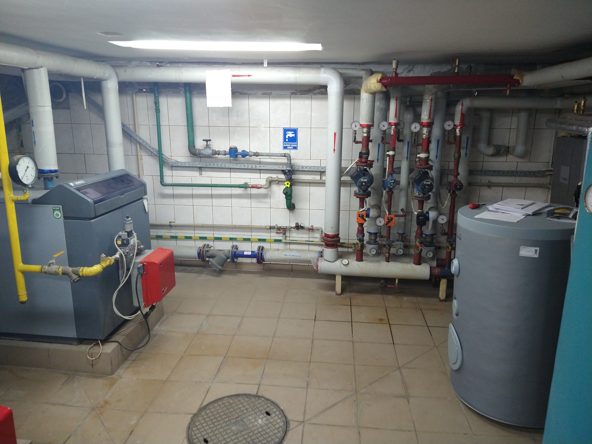 Kotłownia z piecem, zbiornikiem akumulacyjnym i rozbudowaną instalacją rur z zaworami i manometrami, widoczne płytki ceramiczne na ścianach i podłodze.