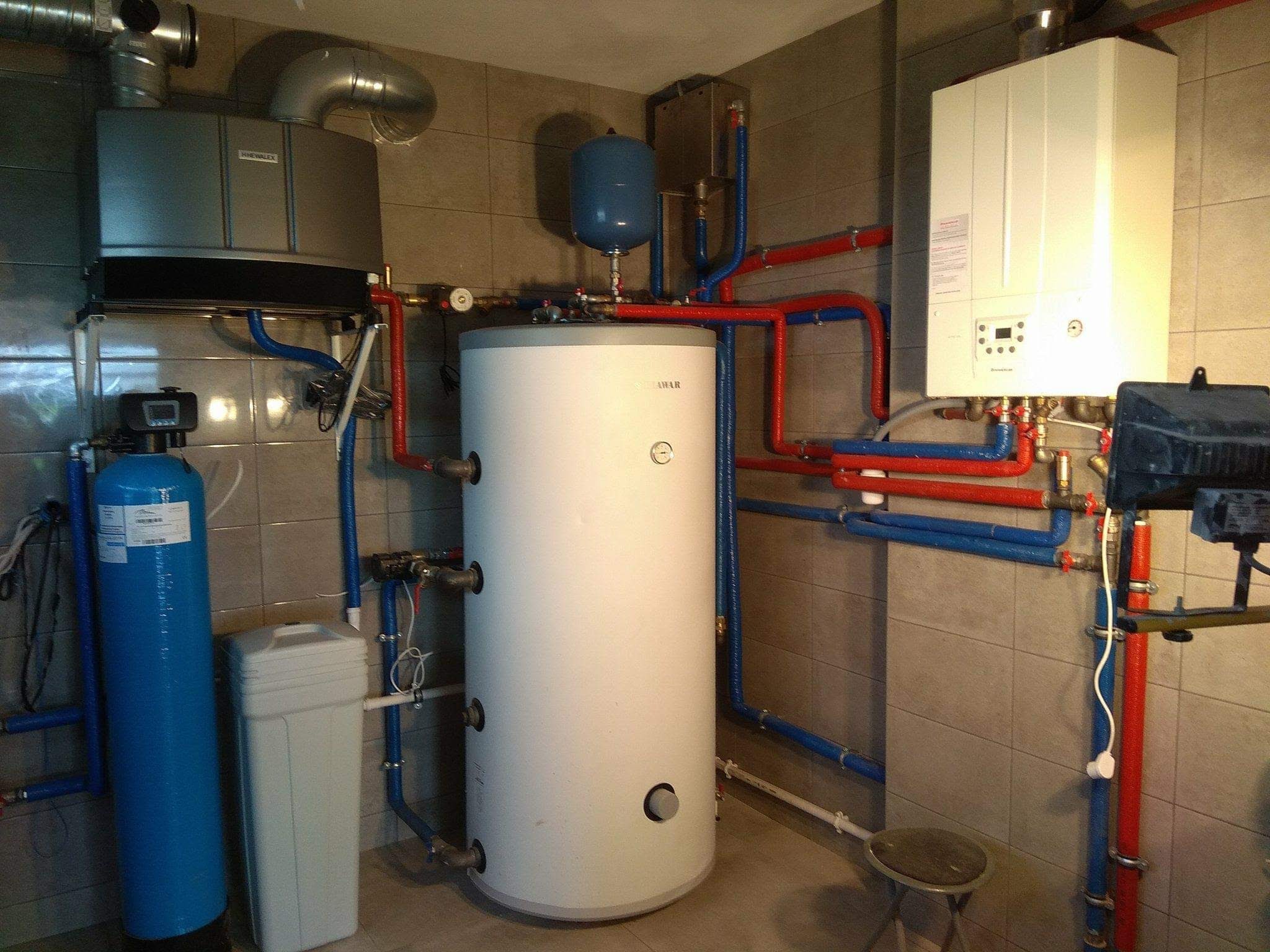 Kotłownia z piecem gazowym, zasobnikiem ciepłej wody, filtrami wody i rurami w kolorach niebieskim i czerwonym, zainstalowana w pomieszczeniu wyłożonym płytkami ceramicznymi.