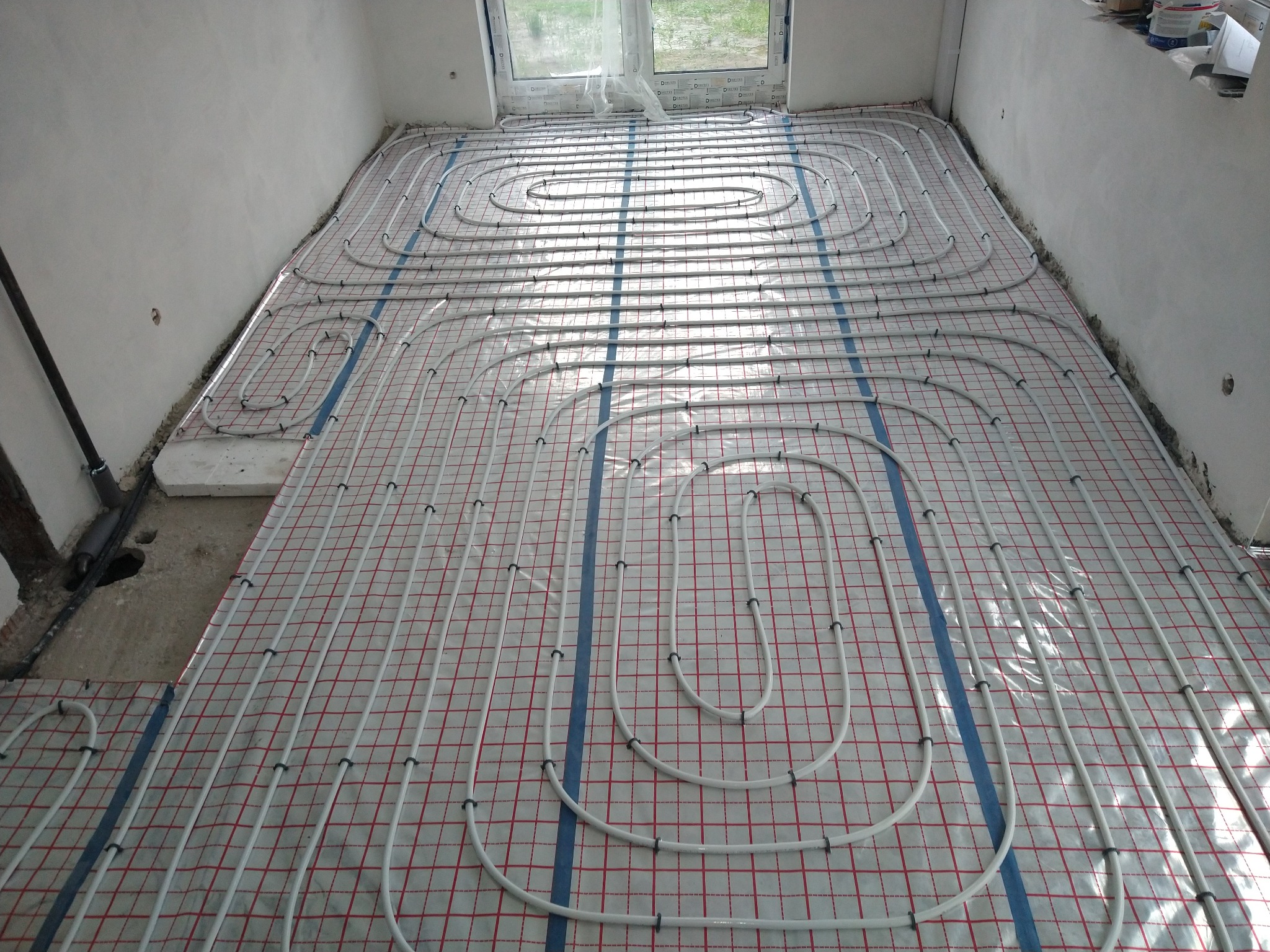 Białe rury ogrzewania podłogowego ułożone na folii z czerwoną kratką, częściowo przykryte betonem, widoczne rury wystające ze ściany, okno w tle.