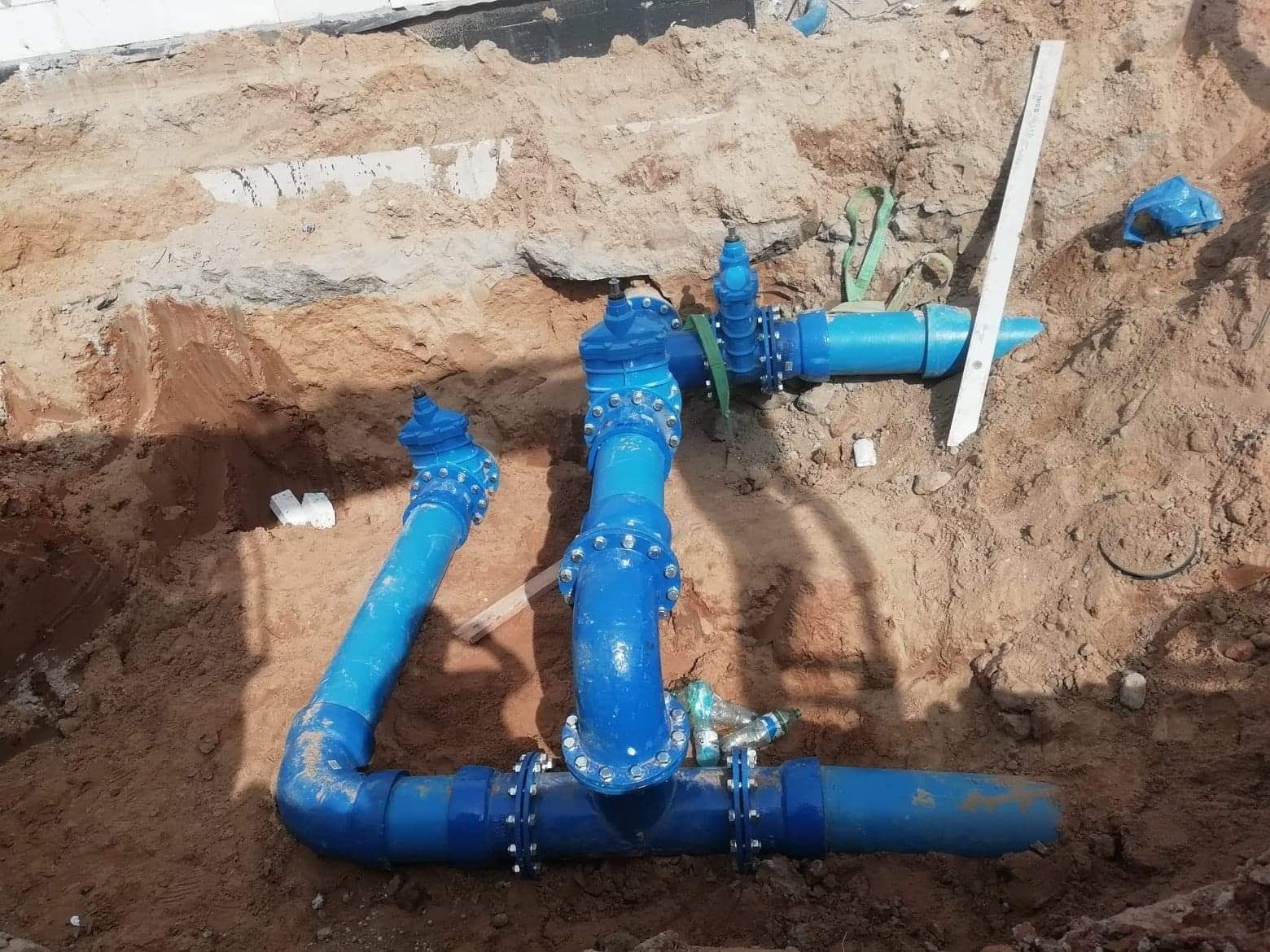 Błękitne rury wodociągowe z zaworami, połączone śrubami, ułożone w wykopie ziemnym, widok z góry na instalację w trakcie budowy lub naprawy.