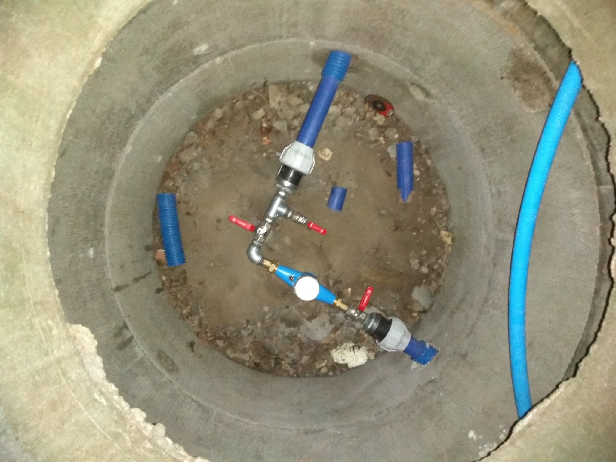 Wnętrze betonowej studni z nowo zainstalowanym systemem rur z zaworami, fragmentem niebieskiej rury i elementami montażowymi, widoczne ślady ziemi i gruzu na dnie studni.