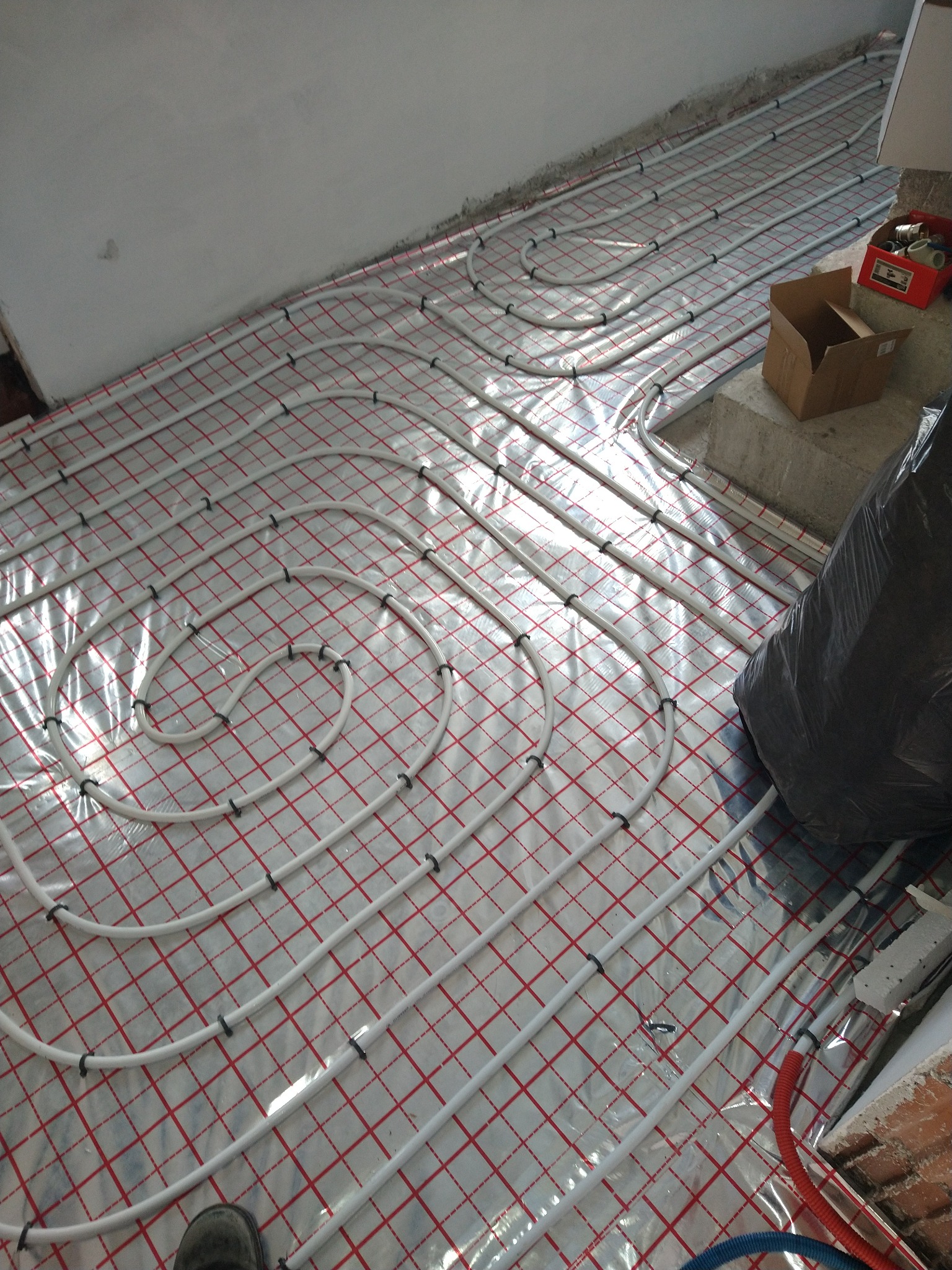 Białe rury ogrzewania podłogowego ułożone spiralnie na folii z czerwoną kratką, widoczny fragment buta pracownika w rogu kadru, obok kartonowe pudełka i skrzynka z elementami instalacyjnymi.