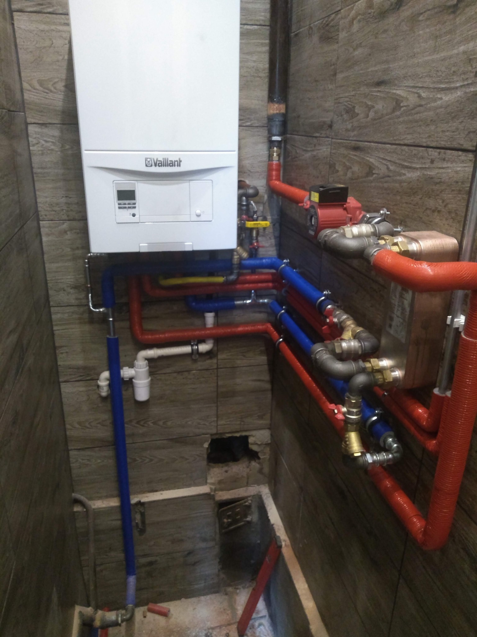 Biały piec grzewczy Vaillant z podłączonymi kolorowymi rurami (czerwone, niebieskie, żółte) i pompą obiegową na tle drewnopodobnych płytek w Tomaszowie Mazowieckim.