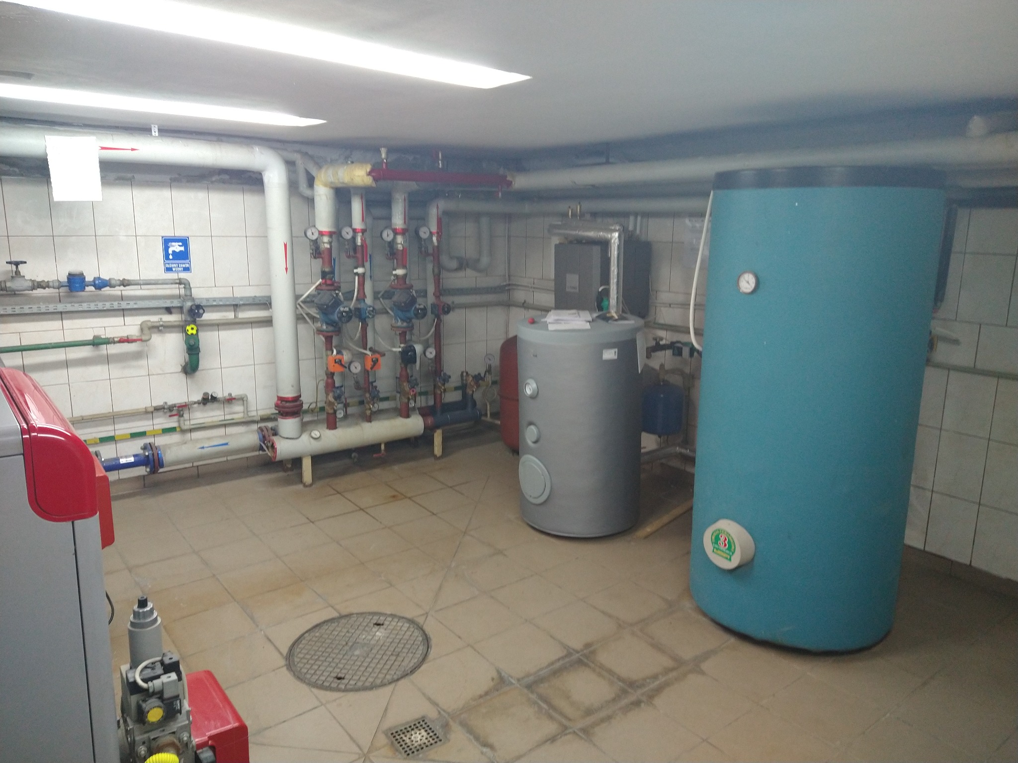 Kotłownia z rozbudowaną instalacją grzewczą, widoczne rury, zawory, zbiorniki buforowe i piec z czerwonymi elementami obudowy.