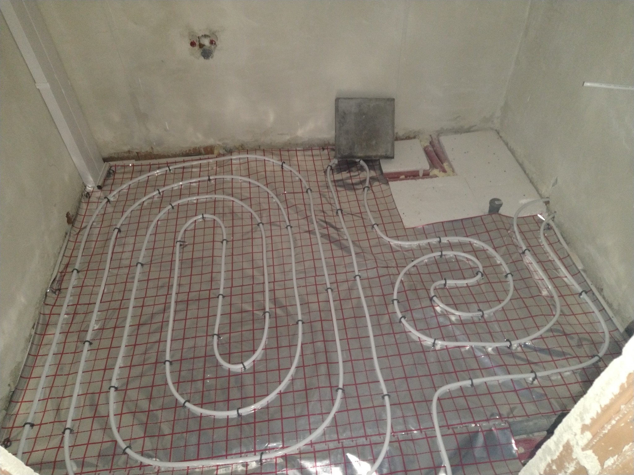 Montaż ogrzewania podłogowego: białe rury ułożone na czerwonej siatce w surowym pomieszczeniu przed wylewką.