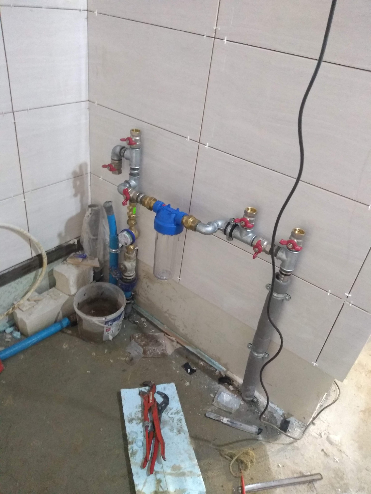 Nowe przyłącze wodne z filtrem i zaworami, widoczne narzędzia na podłodze w trakcie montażu, płytki na ścianach z krzyżykami dystansowymi.