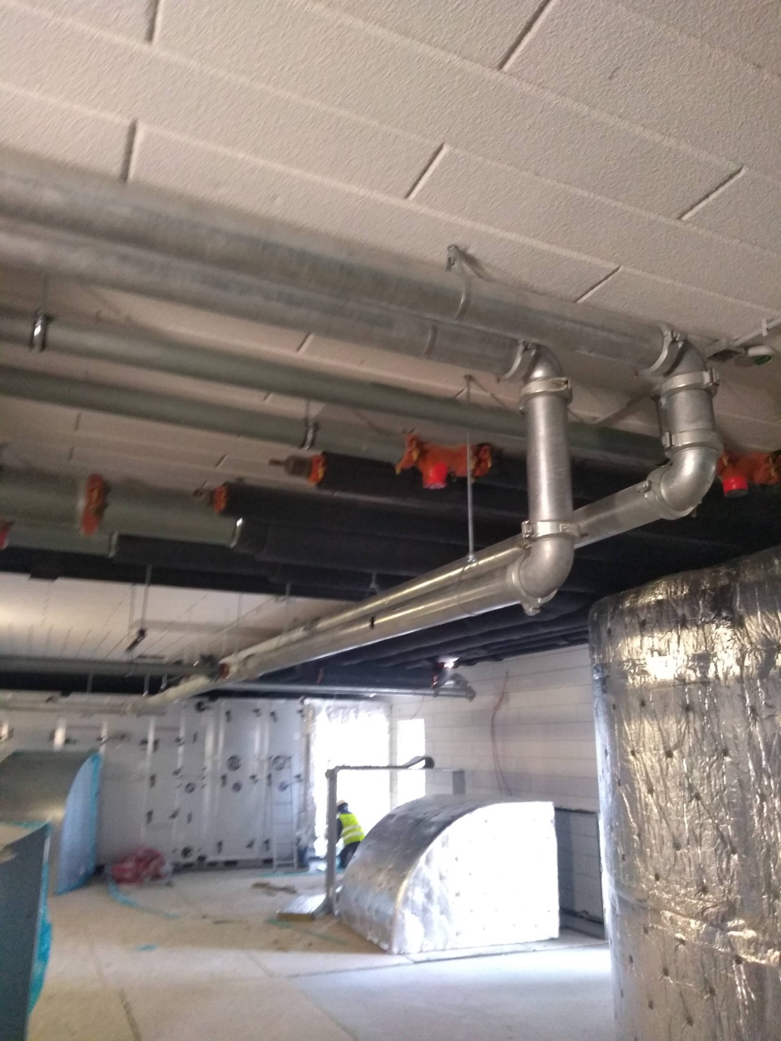 Szare metalowe rury instalacji pod sufitem, widoczne także elementy instalacji przeciwpożarowej w kolorze czerwonym, w tle duże zbiorniki, osoba w kamizelce odblaskowej i drabina.