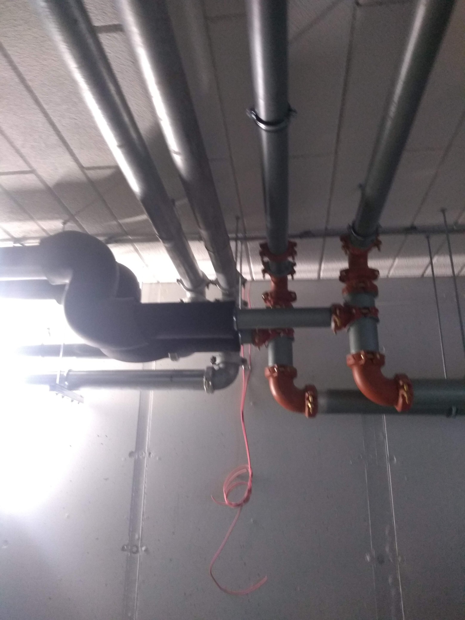 Szare rury pod sufitem z czerwonymi złączkami, widoczne kable i konstrukcja sufitu podwieszanego.