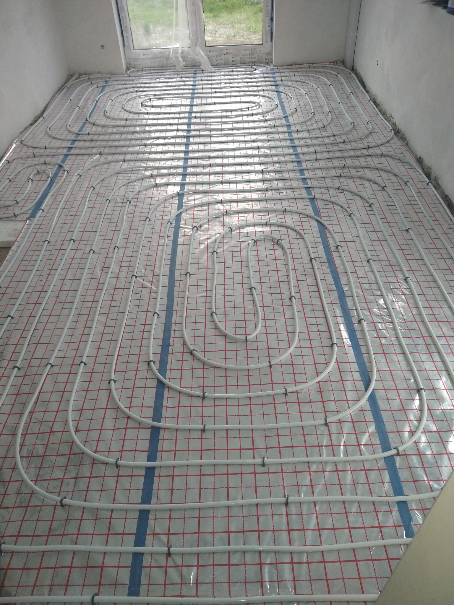 Białe rury ogrzewania podłogowego ułożone na folii z czerwoną siatką w surowym pomieszczeniu z oknem.