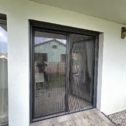 Zewnętrzne drzwi balkonowe z rozsuwaną moskitierą plisowaną, widok z zewnątrz na taras z szarymi płytkami, odbicie fotografa w szybie.