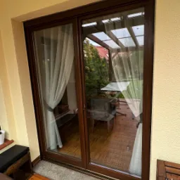 Zasłonięte, przesuwne okno balkonowe z brązową ramą, widok na zadaszony taras z meblami ogrodowymi i zielenią. Na pierwszym planie drewniana podłoga balkonu.