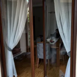 Rozsuwane okno balkonowe z brązowymi ramami, częściowo otwarte, z widocznymi białymi zasłonami i wnętrzem jadalni z osobą siedzącą przy stole.