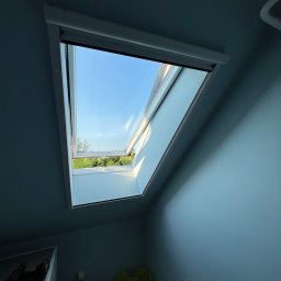 Vladyslav Olefirov - Otwarty okno dachowe z widokiem na błękitne niebo i zieleń drzew, z częściowo zasłoniętą roletą i fragmentem pomieszczenia z pralką i koszem na pranie.