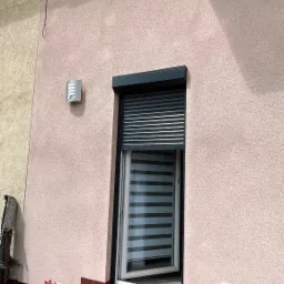 Zewnętrzna roleta częściowo opuszczona na oknie, widok na roletę wewnętrzną, na progu leży pies rasy sznaucer miniaturowy, w doniczkach czerwone pelargonie.