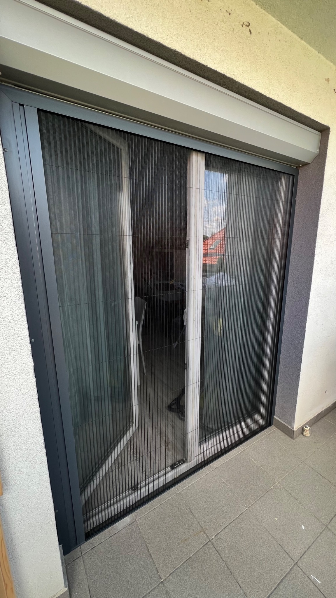 Zamontowana plisowana moskitiera w kolorze grafitowym na balkonie, widoczne otwarte skrzydło drzwi balkonowych, podłoga wyłożona szarymi płytkami.