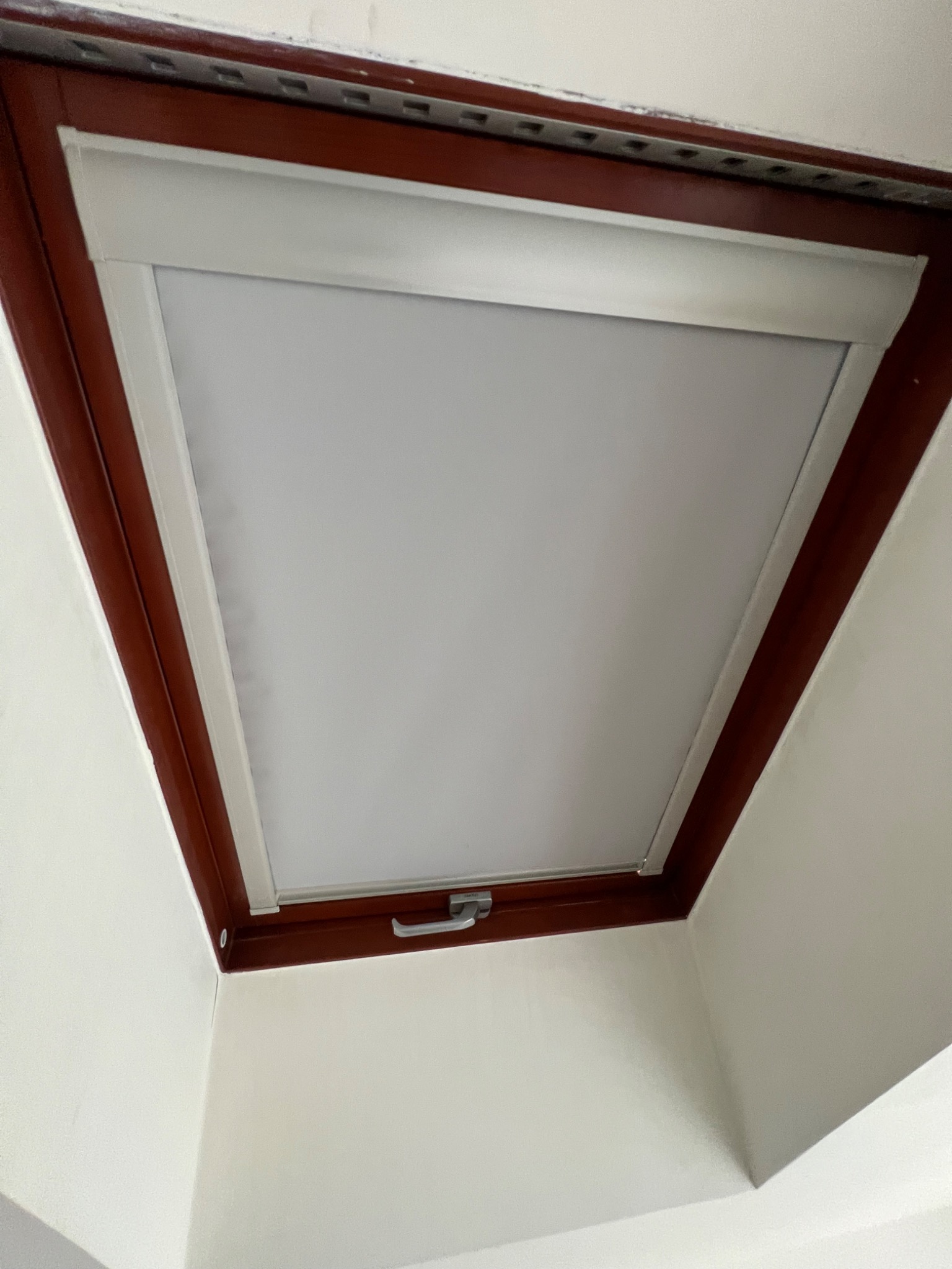 Okno dachowe z wewnętrzną roletą zwiniętą, widok od dołu, brązowa rama okienna, biała ściana.