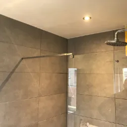 Szklana kabina prysznicowa z chromowanym uchwytem i deszczownicą zamontowana na ścianie z szarych płytek, widoczne odbicie osoby w szkle i okrągły punktowy reflektor w suficie.