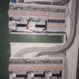 Rozdzielnica elektryczna z licznymi zabezpieczeniami i przewodami, widoczne bezpieczniki i kable w peszlach, całość zamontowana wewnątrz białej obudowy.