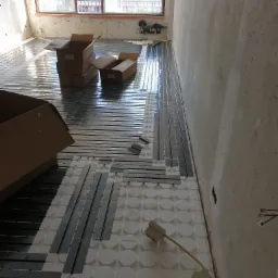 Montaż ogrzewania podłogowego: biała izolacja z okrągłymi wytłoczeniami i czarne rurki grzewcze ułożone w surowym pomieszczeniu z widocznym oknem i kartonami.