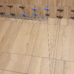 Ułożone płytki imitujące drewno na podłodze i ścianach łazienki, z widocznym odpływem liniowym i elementami poziomującymi płytki w kolorze niebieskim i czarnym.