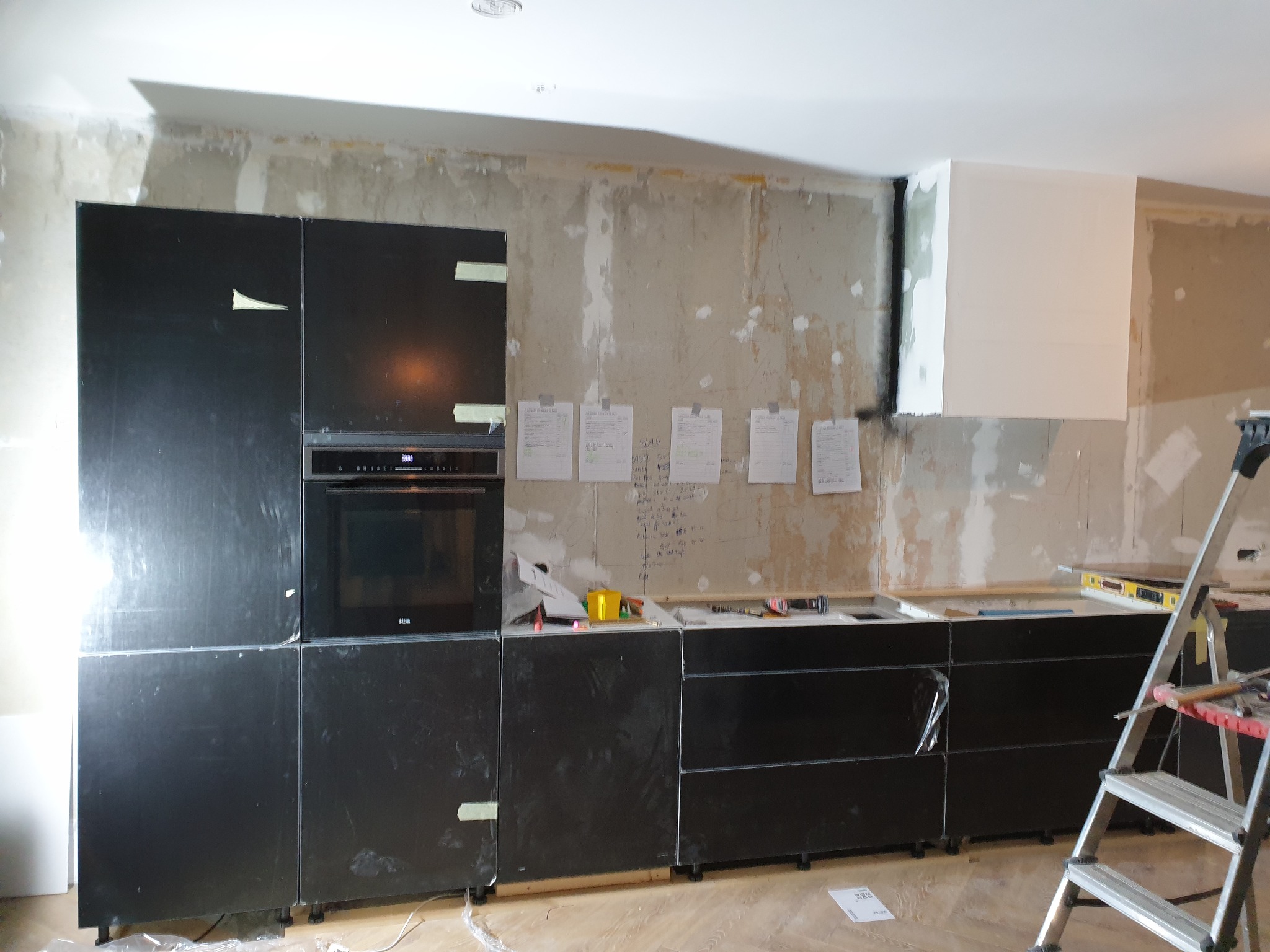 Montaż czarnych szafek kuchennych z piekarnikiem w surowym wnętrzu, ściany w trakcie wykończenia, drabina aluminiowa w prawym rogu, notatki na ścianie.