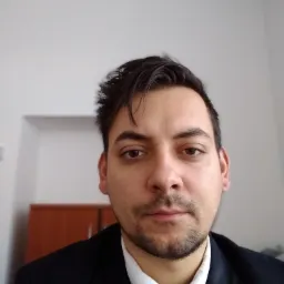 Portret mężczyzny w czarnym garniturze i krawacie, z lekkim zarostem, na tle białej ściany z widocznym fragmentem drewnianego mebla.