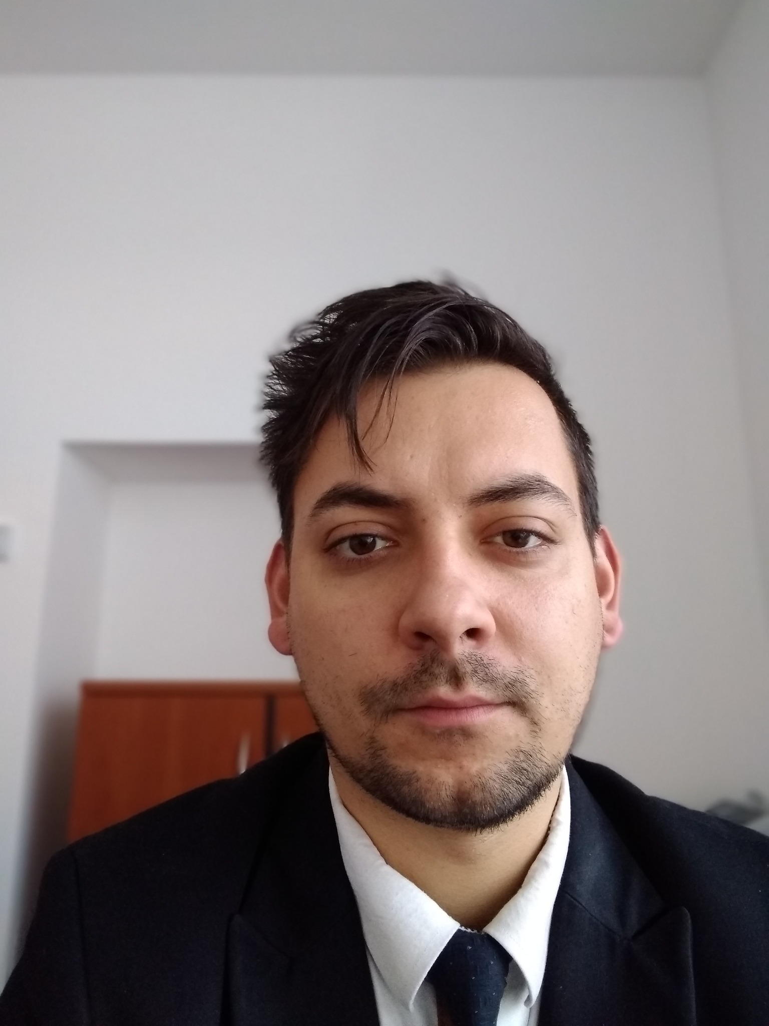 Portret mężczyzny w czarnym garniturze i krawacie, z lekkim zarostem, na tle białej ściany z widocznym fragmentem drewnianego mebla.