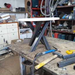 Stół z białym blatem i metalową podstawą w kształcie litery X, spawarka i narzędzia na warsztatowym stole w tle.