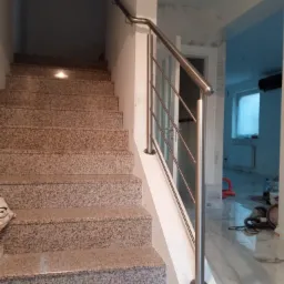 Schody z lastryko z nowoczesną poręczą ze stali nierdzewnej z pionowymi słupkami, widok z dołu schodów, w tle pomieszczenie w trakcie remontu.