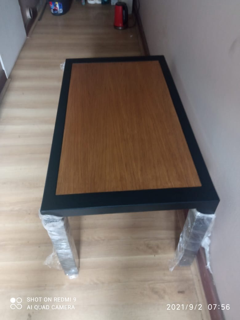 Stolik kawowy z drewnianym blatem i metalową ramą, z nogami zabezpieczonymi folią ochronną, widoczne niedoskonałości podłogi.