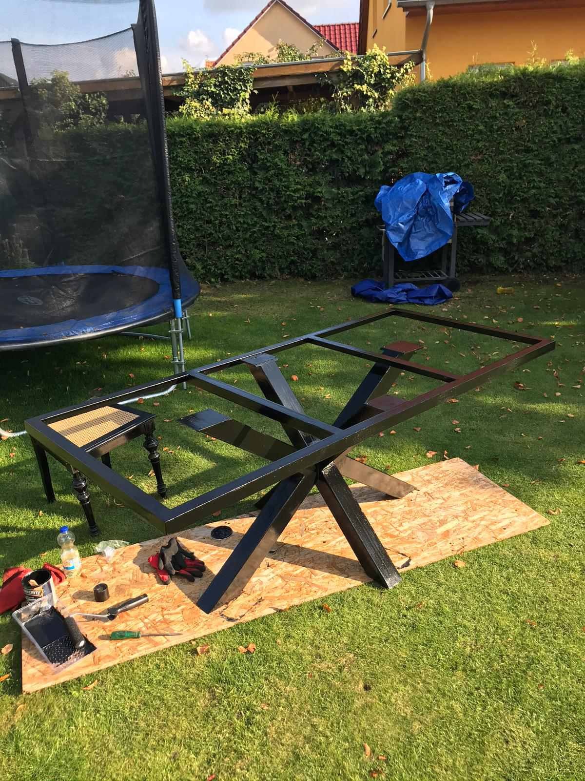 Metalowa konstrukcja stołu w trakcie malowania na zewnątrz, widoczne narzędzia i akcesoria malarskie, trawnik w tle.